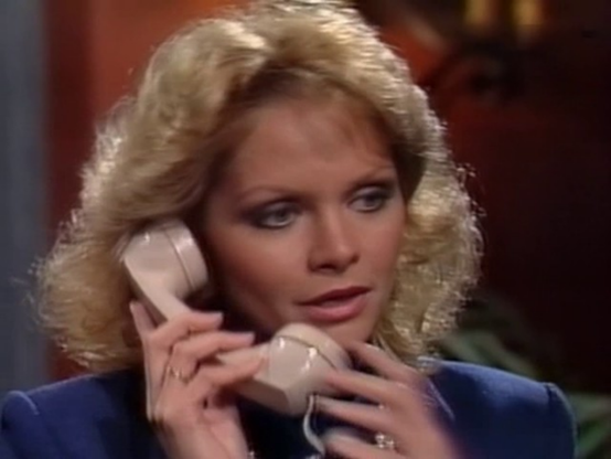 Marcy is in an interior location with an indistinct background.  Her head and shoulders are visible, she is wearing a blue jacket.  She has a phone receiver to one ear, she is holding it with both hands.  She seems deep in conversation.  Her blond hair is wavy and layered, down to her shoulders.