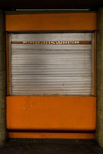 color photograph, portrait format. a closed ticket counter with lowered shutters; half legible in a gap: tickets. silver shutters, orange counter.