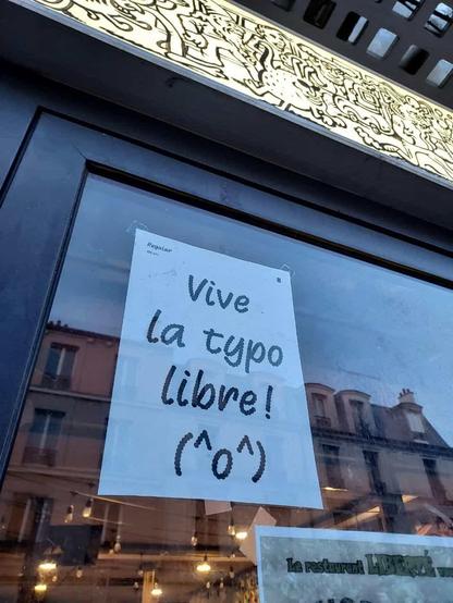 Une photo de la devanture d'un bar avec une affiche typographique en noir et blanc visible à travers la vitre qui dit "vive la typo libre !" avec un petit kaomoji