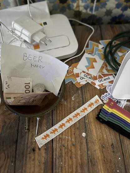 a little bookmark with some two-humped camels cross-stitched in bright orange. also nearby are some OCaml and MirageOS stickers, a piece of network gear, and a bowl with some Moroccan dirham and a note that says “BEER MONEY”