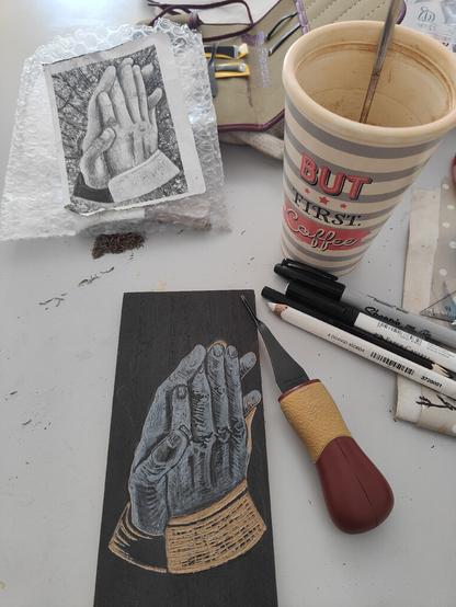 Workstation with pens and tools for carving the block. On the dark surface is the white sketch and some areas are already carved away. My coffee cup is also shown. A flipped black and white print of my painting is propped on the tool case for easy reference.