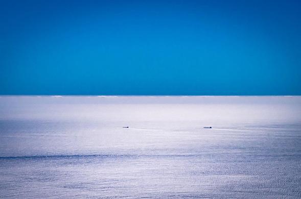 The image shows a calm ocean with a clear blue sky. There are two small ships in the distance on the horizon. The overall scene is tranquil and expansive.