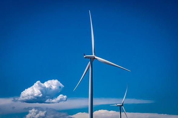 The image shows wind turbines against a clear blue sky with some clouds. Wind turbines are used to generate electricity from wind energy.