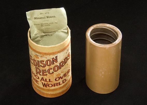 The image features a vintage Edison Records cylinder and its protective sleeve. The cylinder is cylindrical with a light brown color and a label that reads "EDISON RECORDS WHO ALL OVER THE WORLD." The label is ornate, with decorative elements and text in a serif font. The top of the cylinder is open, revealing a green paper insert with the text "No. 4705 Minstrel Record. Three Minutes with the Minstrels." The insert is partially pulled out, showing the text clearly. Next to the cylinder is a matching protective sleeve, also light brown, with a threaded top, indicating it was designed to fit over the cylinder. The background is black, providing a stark contrast to the objects.

Provided by @altbot, generated privately and locally using Ovis2-8B