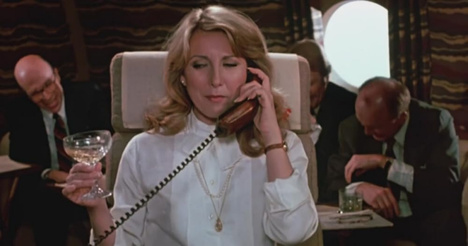 The late great Teri Garr in a still from the movie, "Mr. Mom."  She seems to be in an office setting, sitting in a high backed off white chair.  Two men are working in the background behind her.  She is shown from the waist up, wearing a white blouse.  She has a champagne class in one hand and a phone receiver up to her ear in the other.  Her blond hair is flipped at her shoulders.