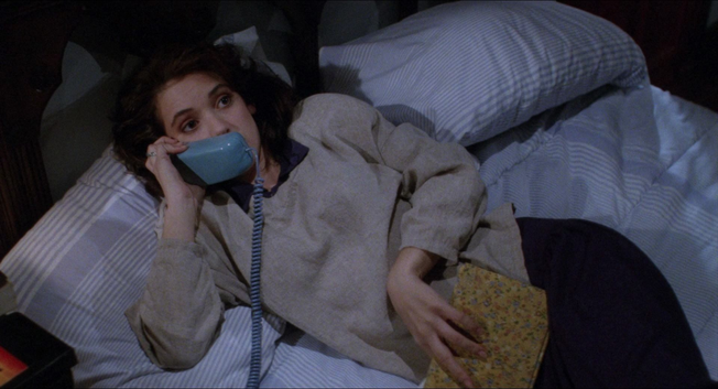 Winona Ryder in a still from the movie "Heathers."  She is lying on her side on a bed.  She is wearing an off white sweater and black pants.  The bed has light blue sheets and two pillows.  She is propped up on one arm, holding a large blue phone receiver.