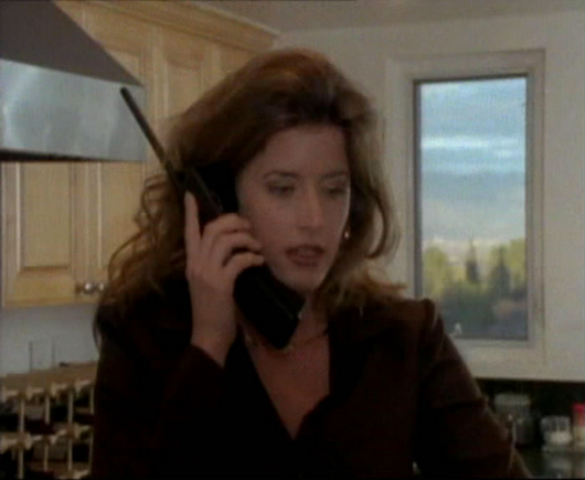 Landon Hall in a still from the 1997 movie, "The Escort" is in an interior location, with a window in the background behind her.  She is dressed in a black jacket with a dark shirt underneath.  She is shown from the waist up.  She has a large, black cordless phone in her hand up to her ear with the antenna extended.  She looks like she is concentrating on the phone conversation.  Her dark curly hair falls down around her shoulders.