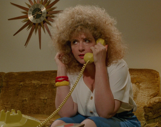 Caroline Aaron in a still from the movie, "Edward Scissorhands."  She is sitting on a brown couch in a room decorated in the style of the 1960s, there is a large starburst clock on the wall behind her.  She is wearing a white blouse and a blue skirt and has a multicolor bracelet.  There is a mustard yellow phone base in the foreground and she has the receiver up to her ear.  Her strawberry blond hair is permed into a huge afro.