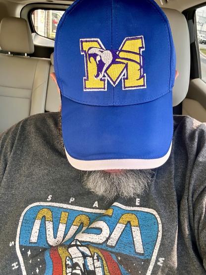 Image of a person sitting in a car, wearing a blue cap with an "M" logo and a gray NASA-themed t-shirt. The focus is on the cap and shirt, with part of the vehicle interior visible.