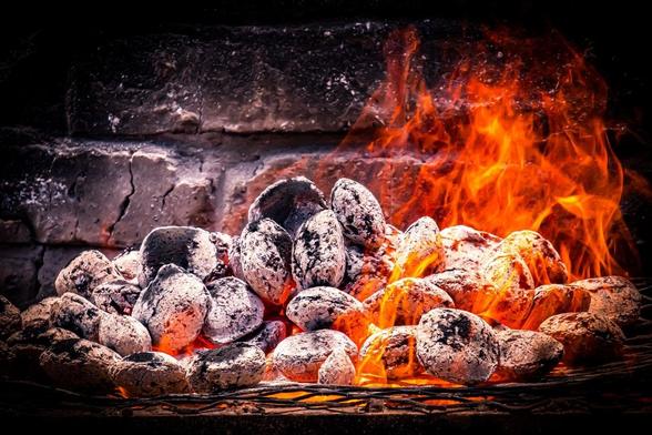 This image shows a pile of burning charcoal briquettes with flames rising from them. They are placed on a wire grate with a brick background, likely indicating a barbecue or a fireplace setting. The briquettes are partially covered in ash, indicating they are actively burning.