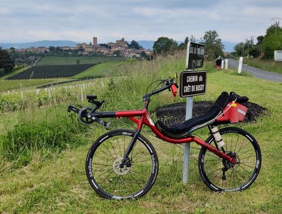 Vélo couché rouge high racer de Metabike. Adossé contre un poteau dans un champs. Au fond on voit le village d'Oingt qui se détache sur les crêtes du Beaujolais.