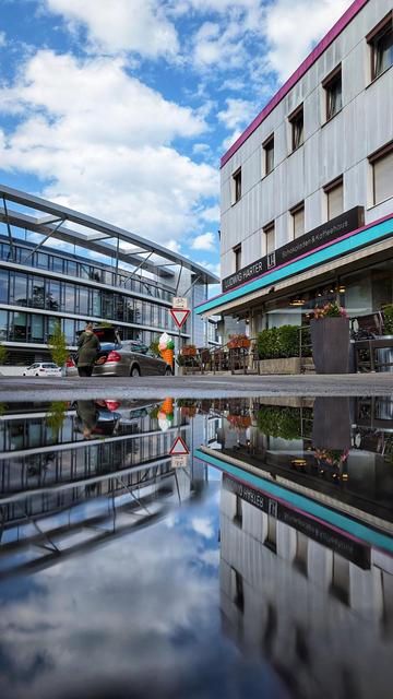 Das Bild zeigt eine Straßenszene mit einem modernen Gebäude im Hintergrund und einem älteren Gebäude im Vordergrund. Das ältere Gebäude hat ein Schild mit der Aufschrift „LUDWIG HARTER“ und „Schokoladen & Kaffeehaus“. Vor dem Gebäude befinden sich Tische und Stühle, die zu einem Café gehören. Auf der Straße steht ein Auto mit geöffnetem Kofferraum, daneben eine Person. Ein Verkehrsschild mit einem Fahrrad-Symbol und einem Vorfahrt-gewähren-Zeichen ist ebenfalls sichtbar. Eine interessante Besonderheit des Bildes ist die Spiegelung der gesamten Szene in einer Pfütze auf der Straße