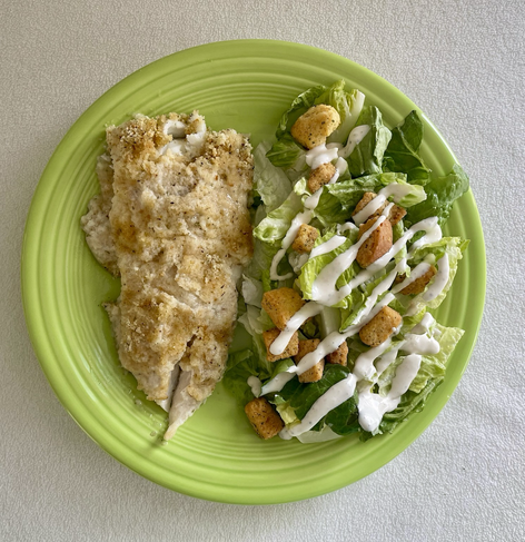 On a light colored green plate sits dinner. On the left baked haddock. On the right a Caesar Caesar salad salad. There are croutons, multi shades of green lettuce, and white drizzled dressing.
