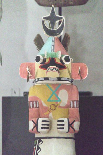 Nom : Kachina (détail)
Auteur : Culture hopi (Arizona, États-Unis)
Date : XXI
Type : Artisanat sur bois peint, découpé et collé
Objet : Poupée
Source : Musée laïque d’Art Sacré du Gard
Cote/Inv. : CD012.4.1
Document : ORAEDES (2024)
Commentaire : Les kashinas figurent les forces animiques et spirituelles liées à toute chose dans le cosmos, d'un objet matériel précis à un concept abstrait et général. Ces entités visitent périodiquement les humains afin de les épauler dans leurs activités quotidiennes et religieuses; eux-mêmes vivent en société. Fabriquées dans des racines de peuplier, ces poupées sont destinées aux enfants Hopis et Zuñis afin de les familiariser avec l'apparence de ces entités et de fixer leur imagination. Elles sont plus spécialement confiées aux filles. En effet, outre le fait d'avoir adopté le système des kachinas — comme tous les Pueblos — ces peuples ont développé une symbolique et un folklore plus élaboré et pratiquent plus spécialement les rites en rapport. Le terme 'kachina' désigne tout à la fois l'entité, les danseurs masqués pratiquant le rituel et vêtus pour leur ressembler et enfin les poupées figurant simultanément entités et danseurs. Ces cérémonies religieuses ont lieu plusieurs fois au cours de l'année. Les couleurs utilisées pour les vêtements et les poupées sont naturellement symboliques, en rapport avec le système de directions spatiales des pueblos. [suite sur Oraedes]