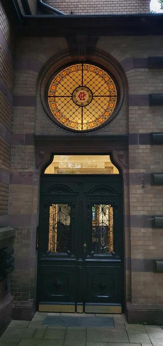 above a black wooden doorway there is an ornamented round glass window with a red "R" on orange glass