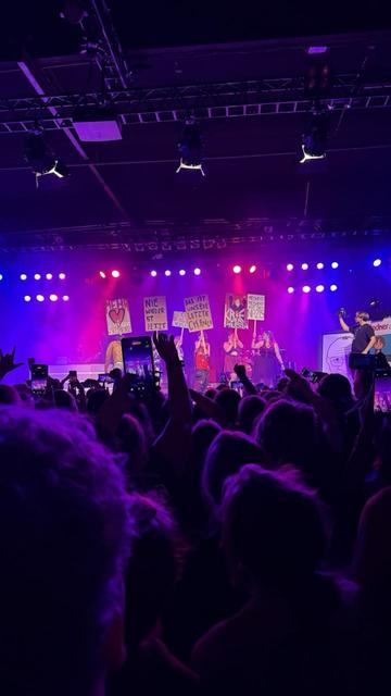 Das Bild zeigt eine Konzertszene (Yu Konzert in Köln) in einer Halle mit einer großen Menschenmenge im Vordergrund. Viele Personen im Publikum halten ihre Smartphones hoch und filmen oder fotografieren die Bühne. Die Bühne ist in buntes Licht getaucht – vor allem Lila-, Blau- und Rottöne dominieren.

Auf der Bühne stehen mehrere Menschen mit Plakaten in der Hand. Die Botschaften auf den Plakaten sind politisch und gesellschaftskritisch, unter anderem steht dort:
	•	„Mehr Liebe, weniger Sch***e“
	•	„Nie wieder ist jetzt“
	•	„Das ist unsere letzte Chance“
	•	„Krieg den Palästen“
	•	„Kein Moshpit für Nazis“

Links und rechts auf der Bühne sind Zeichnungen oder Banner mit weiteren Botschaften oder Illustrationen zu sehen. Die Stimmung wirkt ausgelassen, engagiert und solidarisch – es scheint sich um ein Konzert mit politischer Botschaft zu handeln.