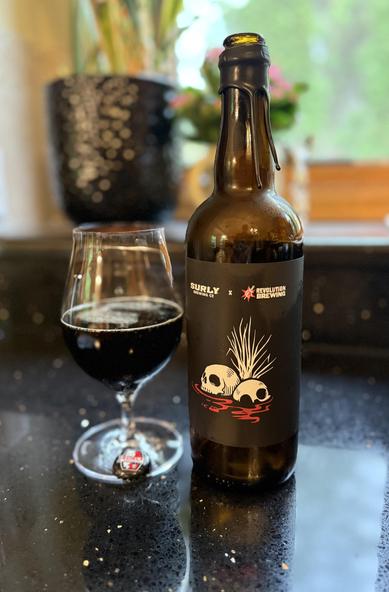 A snifter glass of dark beer next to a tall brown bottle with a black label and two white skulls sinking into a red surface