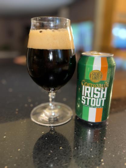 A tall snifter glass of dark beer next to a 12 ounce can in vertical stripes of dark green, light green, white and orange