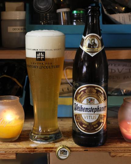 A pilsner glass filled with light amber fluid and topped by a thick layer of white foam stands next to a dark brown bottle of beer. The label on the bottle is a white oval with a gold and maroon design, and reads “Weihenstephaner Vitus Weizenbock.”