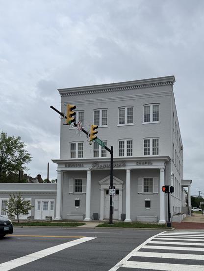 The image shows a three-story building with a gray facade and white trim, prominently labeled "MEMORIAL Demaine's CHAPEL" on the front. The building features a symmetrical design with evenly spaced windows on each floor and a central entrance flanked by white columns. The roofline is adorned with decorative molding, and the building is situated at an intersection, as indicated by the traffic lights and street signs. The street signs include a green street name sign reading "Garrison St." and a no left turn sign. A pedestrian crossing is visible in the foreground, with a red pedestrian signal light. The sky is overcast, and the overall color palette is muted, with the gray of the building contrasting against the white trim and the darker tones of the street and vehicles.

Provided by @altbot@fuzzies.wtf, generated privately and locally using Ovis2-8B

🌱 Energy used: 0.191 Wh