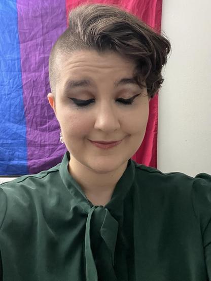 selfie of a young white person with short brown hair (me). I'm wearing a full face of makeup and a green shirt with a bow at the neck. I’m standing in front of a bi flag.
