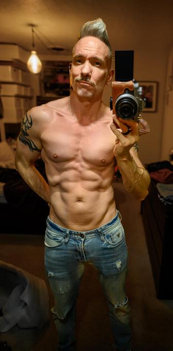 Shirtless bedroom mirror selfie standing wearing only ripped up blue jeans, holding up my Nikon z fc in my right hand, taking the picture, with my left hand pulled behind my back, light shining down from above highlighting   my swooped up hair, and the usual muscly stuff like shoulders, pecs, abs and cum gutters, and of course the smirk™