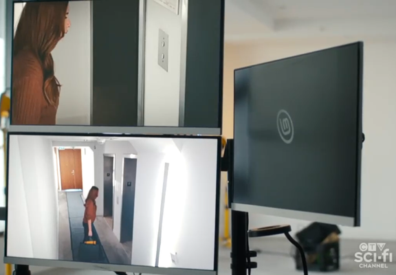 Screen cap from TV show showing some cctv and computer monitors.  The one on the right appears to be sleeping behind a Linux Mint logo.