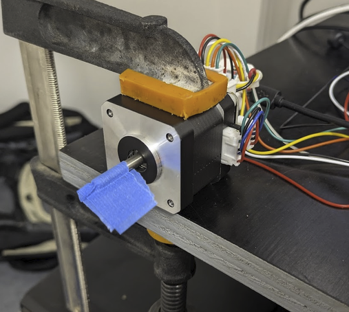 A photo of a NEMA17 stepper motor clamped to a benchtop. There is a flag of blue tape attached to the stepper shaft. Behind the stepper is a tangle of multicoloured wires.