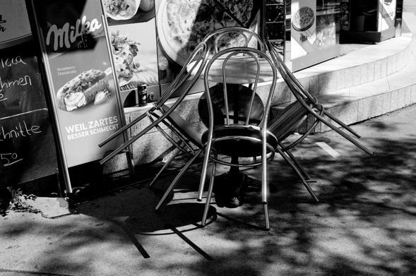 The black and white image shows a group of metal chairs stacked upside down on top of each other around a small outdoor café table. In the background, there are colorful posters advertising food, including a Milka strawberry cheesecake ice cream bar. A shadowy tree canopy dappled across the pavement adds texture to the urban sidewalk setting.