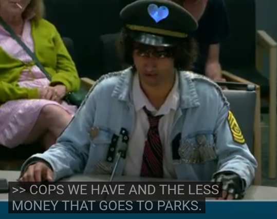 at 4h22 in the video, wearing a michael jackson getup with a mirrored heart as a badge on a police hat, saying "the more cops we have, the less money that goes to parks"