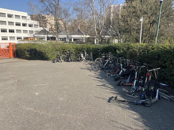 Vue d’une place à l’entrée d’un collectif avec des trottinettes accrochées en vrac sur des arceaux vélo, on en distingue trois dans le tas