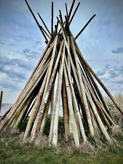 This image features a large, cone-shaped structure made from many long wooden poles or logs arranged leaning together at the top, reminiscent of a teepee construction. It's located in a natural setting with visible grass in the foreground and a body of water and overcast sky in the background. The structure likely serves a decorative or functional role in this outdoor setting, possibly used for shelter or as a visual accent in the landscape.