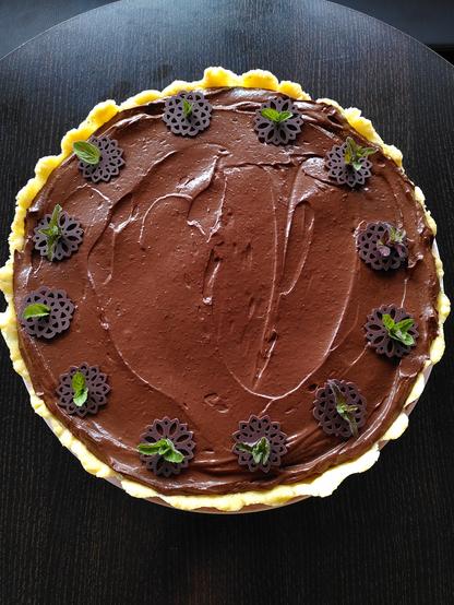 Eine große Schoko-Minz-Mousse-Tarte. Die Schokoladencrème glänzt und macht Appetit. Dekoriert ist die Tarte mit filigranen Schokoladenplättchen und frischen Minzblättchen.
Der Mürbeteigrand ist ebenfalls zu erkennen und bildet einen schönen Kontrast zur dunkelbraunen Füllung.