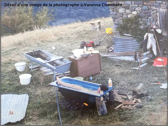Photo. Sur un terrain en pente herbeuse près d’un bâtiment en pierre. Une femme est allongée dans une baignoire bleue, remplie d’eau, posée sur deux troncs de bois, avec un feu allumé en dessous. Autour de la baignoire, plusieurs objets sont disposés : planches de bois, bouteilles en plastique, bassines, ustensiles de cuisine, une bassine métallique sur pieds, un meuble en bois servant de plan de travail, des seaux, des vêtements suspendus et des éléments de bricolage. À l’arrière-plan, un paysage vallonné et boisé.