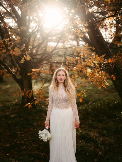 This image by Hamburg based European elopement and wedding photographer Sturmsucht captures a person standing in a forest during autumn. They have long, wavy blonde hair and are wearing an elegant white outfit consisting of a delicate, sheer top with intricate beaded or lace patterns and a flowing, pleated white skirt. They're holding a small bouquet of white flowers in one hand.

The setting is particularly striking - they're surrounded by trees with vibrant orange and golden autumn leaves. Sunlight filters through the branches from above, creating a warm, golden glow that illuminates the scene and creates a halo effect around the subject. The forest floor is covered with grass and fallen leaves, enhancing the autumn atmosphere.

The overall mood is serene and ethereal, with the contrast between the white outfit and the warm autumn colors creating a dreamlike quality. The lighting is soft and warm, suggesting it might be taken during the "golden hour" near sunset or sunrise.