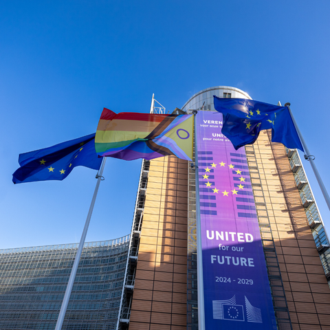 The progressive flag flies between two European Union flags in front of the European Commission headquarters in Brussels. Behind them, a large purple banner on the Berlaymont building reads “United for our Future 2024–2029” with a circle of yellow stars forming the EU symbol.