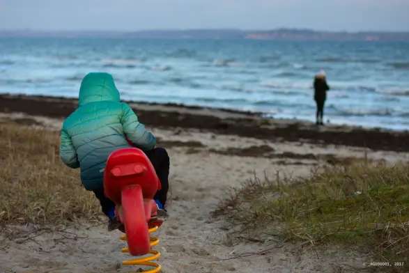An einem Sandstrand steht ein rotes Kindermotorrad auf einer gelben Feder. Man sieht das Spielzeug von hinten. Auf dem Motorrad sitzt ein Kind, das sich in die Kurve legt. 
Im Hintergrund ist die Flensburger Förde zu sehen. Die Ostsee ist ein bisschen kabbelig, am Ufer steht eine Person. Am Horizont ist Dänemark zu erkennen.