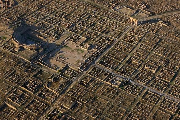 An aerial view of the extensive ruins of a Roman town, showing a well-preserved grid layout with stone foundations, streets, and colonnades. Visible landmarks include a large amphitheater, a central forum surrounded by columns, and monumental arches, all set within a geometric network of buildings and thoroughfares.