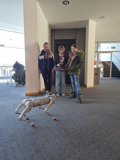 Credit: Hochschule Hof  
Alt-Text: Vier Kinder stehen in einem Innenraum auf einem Teppichboden und beobachten einen vierbeinigen Roboter mit der Aufschrift „GO1“. Eines der Kinder steuert den Roboter mit einer Fernbedienung. Im Hintergrund sitzt eine weitere Person an einem Tisch nahe einem Fenster./ Four children are standing indoors on a carpeted floor, observing a four-legged robot labeled "GO1". One of the children is controlling the robot with a remote. In the background, another person is seated at a table near a window.