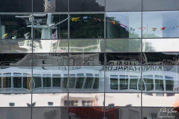 Photo en couleur présentant le reflet, dans la façade vitrée d'un immeuble de bureau, d'une partie des ponts supérieurs d'un navire sur lequel est hissé le grand pavois.