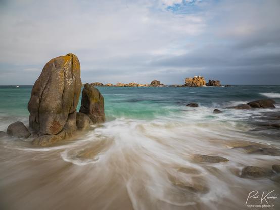 Photo en couleur présentant le reflux, capté en pose longue, de la mer sur le sable d'une plage. Sur cette plage, il y a également des rochers et notamment un petit amoncellement sur la gauche du cadrage avec essentiellement deux gros rochers de forme bien verticale et comme posés sur d'autres plus petits rochers, faisant penser à deux menhirs. À droite de la photo, il y a également des rochers, plus petits et "émergeant" à peine du sable ou de la mer. Sur la ligne d'horizon, d'autres rochers au large, bénéficient d'un rayon de soleil qui les met bien en valeur sous un ciel plutôt nuageux. Au bord de la plage, la mer est de couleur plutôt verte, alors que plus on s'éloigne du rivage, plus elle vire au bleu. Du fait de la captation en pose longue le reflux de la vague précédente, génère de longues traces blanches sur le sable.