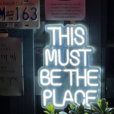 A glowing white neon sign reading 'THIS MUST BE THE PLACE' mounted on a wall, with green plants visible at the bottom. Above the sign is a British Columbia license plate reading 'M 163' from September 1996, and a framed French text about yogurt. The warm, inviting atmosphere suggests a casual bar or gathering space where neighbors might celebrate spring together at outdoor beer tables.