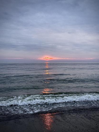 This image captures a serene beach scene at sunset. It features gentle waves coming onto the shore, with the sun partially visible as it sets on the horizon, casting a warm reflection on the water. The sky ranges in color from pale blue to shades of pink and orange near the disappearing sun, creating a peaceful and picturesque setting. The beach appears to be tranquil, without any visible people or significant activity, emphasizing the natural beauty of the seascape.