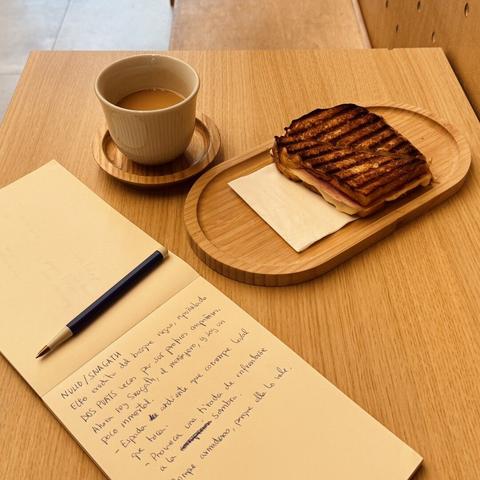 Bodegón de desayuno catalá con mucha base de madera, café en taza SIN ASA, con leche fría y un mixto en pan de molde! Al lado está la libreta con los apuntes de mi personaje.