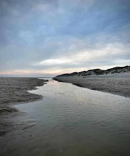 This image captures a serene beach scene with a gentle waterway meandering through the sand. On either side, there are sandy dunes lightly covered in grasses and shrubs, typical of coastal landscapes. The sky has a soft gradient of blues and faint pink, possibly indicating late afternoon or early evening. The tranquility of the scene, along with the natural curvature of the water's path, creates a peaceful, picturesque vista.