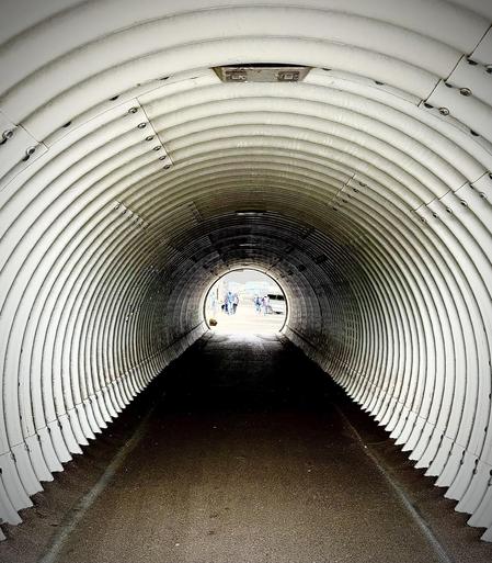 The image depicts a tunnel with a ribbed, corrugated design, possibly made of metal. The tunnel's structure creates a vanishing point effect, focusing attention on the light at the end where several individuals can be seen walking towards and through the tunnel. The perspective provided by the corrugation adds depth to the image, leading the viewer’s eye from the darker interior towards the brightly lit exit.