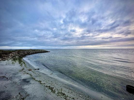 The image shows a tranquil seascape featuring a curved shoreline with a sandy beach on the left side extending towards a stone breakwater or jetty. Gentle waves are lapping against the shore. The sky above is overcast, filled with textured clouds that suggest an early morning or late afternoon light. The calm sea and scattered seaweed along the sand offer a peaceful coastal scene, typical of a quiet, natural setting.