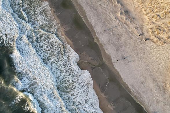 The image shows an aerial view of a beach where the sea meets the shoreline. The waves are creating white foam as they break upon the sandy beach. The shore displays varying shades of sand, from lighter patches to darker, possibly wet areas. There appear to be a few individuals standing on the beach, visible as small figures from this altitude. The textures visible in the sand suggest the presence of ridges or patterns formed by the movement of water. The overall scene captures the dynamic interaction between ocean and land.