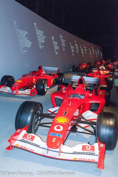 Auf dem Bild sind mehrere  Formel-1-Rennwagen zu  sehen, auf denen die Marke Ferrari deutlich zu sehen ist. Die Autos sind in einer Reihe aufgereiht, wobei das nächstgelegene Auto im Vordergrund die Nummer 1 und das Shell-Logo auf seinem Frontflügel trägt. Die Autos sind mit verschiedenen Sponsorenlogos geschmückt, darunter Vodafone, Bridgestone und Shell. Im Hintergrund ist eine große Wand mit Text zu sehen, der wahrscheinlich einen historischen Kontext oder Informationen über die Autos liefert. Die Beleuchtung ist gedämpft und lenkt die Aufmerksamkeit auf die Autos, und die Gesamtumgebung scheint eine Ausstellung oder ein Museum zu sein. Die Autos sind auf einer ebenen Fläche positioniert, und das Bild fängt die komplizierten Details der Fahrzeuge ein, wie z. B. die aerodynamischen Komponenten und Sponsorenaufkleber.