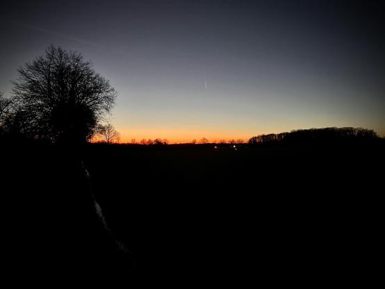 The image captures a striking sunset with a gradient of vibrant oranges fading into deep blues at dusk. Silhouettes of trees outline the horizon, and there is a clear sky with a visible contrail from an airplane. The foreground appears to be a field or open countryside, a small body of water or a wet path reflects the last light on the left side. The scene conveys a tranquil and serene end of the day in a rural setting.